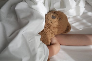 Beyaz çarşafların altında uyuyan, oyuncak ayıya sarılan çocuk. Çocukların uyku zamanı ya da yatma zamanı..