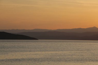 Canlı turuncu günbatımında durgun deniz suyunun arkasındaki dağ tabakalarının silueti.