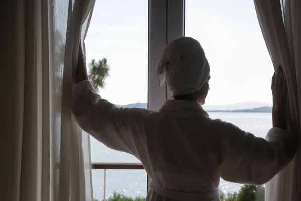 美しい静かな海の景色と朝の部屋でカーテンを開けるバスローブの女性 — ストック写真