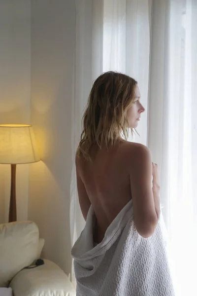 迷人的女人裹着白色的毯子站在一个巨大的窗前 回头看 — 图库照片