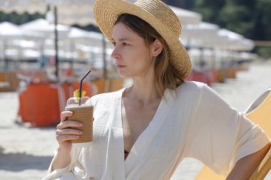 Genç bir kadın plajda gün boyu eğleniyor, güneş sandalyesinde oturuyor ve içecek bir şeyler içiyor.