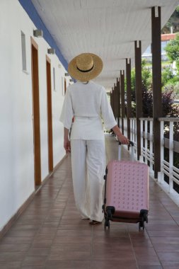 Tekerlekli bavul taşıyan güneş şapkalı bir turist, otel odasına doğru yürüyor. Seyahat, tatil, tatil, yaz tatili, tek başına seyahat kavramı..