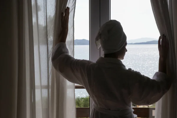 Woman in a bathrobe opening curtains in room in the morning with beautiful peaceful seaview