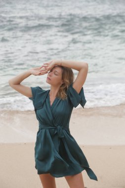 Güzel genç bir kadının portresi. Boş bir kumsalda, şık yeşil ipek elbiseler içinde. Şehirden uzakta huzurlu bir yaz tatili. Zarif ve şık yaz kıyafetleri, bahar-yaz moda trendi.