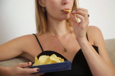 Genç üzgün kadın sağlıksız atıştırmalıklar yiyor, oturma odasında patates cipsi yiyor. Abur cubur ve sağlıksız yeme konsepti.
