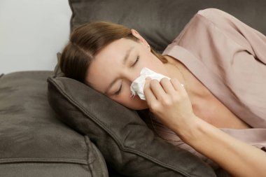 Kanepedeki genç kadın beyaz kağıt mendile burnunu sümkürdü. Alerji belirtileri olan bir kız mendile hapşırıyor. Grip, soğuk algınlığı veya alerji belirtileri