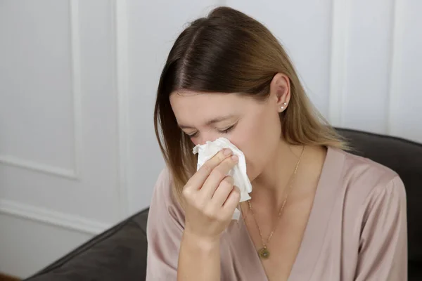 Young woman on the sofa blowing nose into a white paper tissue. Girl with allergy symptoms sneezing into a tissue. Flu, cold or allergy symptom
