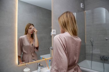 Banyoda aynanın önünde sabah akşam rutinini yapan kadın, kişisel temizlik ve güzellik konsepti.