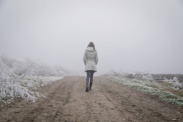 Женщина в белом халате идет по сельской дороге в холодный туманный день. Зимняя погода.