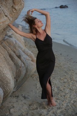 Gecelik siyah uzun elbiseler içinde güzel genç manken, kayalık sahilde moda yaz portresi.