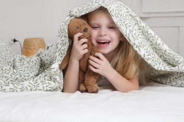 Sevimli küçük anaokulu kızı oyuncak ayısını tutuyor ve yatak odasında battaniyenin altına saklanıyor.