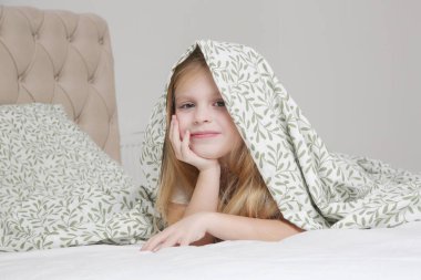 Küçük sevimli anaokulu kızı battaniyenin altında saklanıyor, gülümsüyor ve kameraya bakıyor.