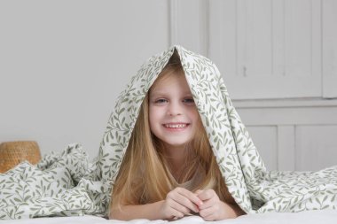 Küçük sevimli anaokulu kızı battaniyenin altında saklanıyor, gülümsüyor ve kameraya bakıyor.