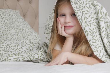 Küçük sevimli anaokulu kızı battaniyenin altında saklanıyor, gülümsüyor ve kameraya bakıyor.