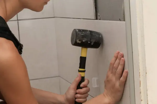Woman tiling bathroom walls, pressing tile into a glue with rubber hammer. Process of installation of tiles in the bathroom step by step. DIY home improvement.