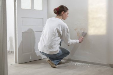 Duvardaki güherçile sorunu. Kadın, sağlam bir yüzeye ulaşana kadar kötü durumdaki boya ve alçıyı kazıyıp çıkarmak için kazıyıcı kullanıyor.. 