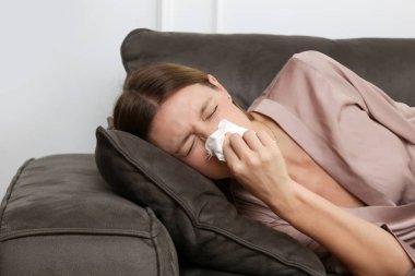 Kanepedeki genç kadın beyaz kağıt mendile burnunu sümkürdü. Alerji belirtileri olan bir kız mendile hapşırıyor. Grip, soğuk algınlığı veya alerji belirtileri