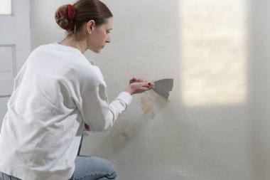 Duvardaki güherçile sorunu. Kadın, sağlam bir yüzeye ulaşana kadar kötü durumdaki boya ve alçıyı kazıyıp çıkarmak için kazıyıcı kullanıyor..