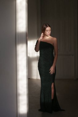 Bir omuz asimetrik elbiseyle parıldayan zarif siyah modelin iç mekan portresi.