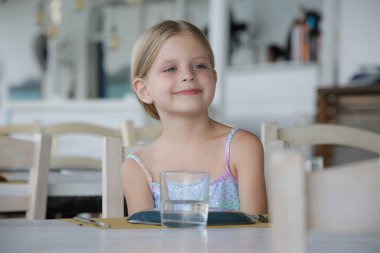 Önünde bir bardak suyla restoran masasında oturan sevimli küçük kızın portresi, sıcak yaz günlerinde susuzluk..