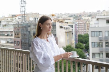 Şehir manzaralı balkonda kahve içen genç bir kadın. Yoğun bir günden önce sakin sakin bir sabah.
