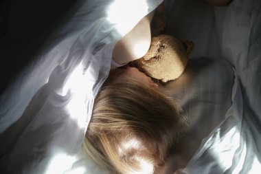 Beyaz çarşafların altında uyuyan, oyuncak ayıya sarılan çocuk. Çocukların uyku zamanı ya da yatma zamanı..