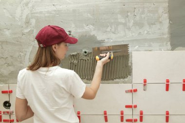 Banyo duvarlarını döşeyen bir kadın fayansa yapıştırıcı sürüyor ve seramik fayansları doğru pozisyona yerleştiriyor. Banyoya fayans döşeme işlemi adım adım yapılıyor. DIY Ev Geliştirme.