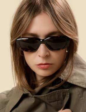 Yeşil trençkotlu, kaplumbağa kabuklu güneş gözlüklü ve perdeli genç kadın portresi.