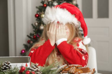 Üzgün, yılbaşı kıyafetli küçük kız ve Noel Baba şapkası masada zencefilli kurabiyelerle oturuyor, kış tatili stresi konsepti.