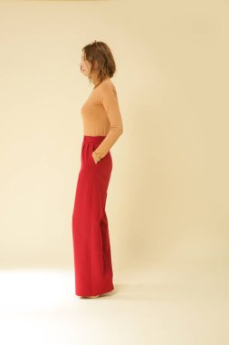 Stüdyo fotoğrafları. Sade güzel kıyafetler giyen, bej balıkçı yaka ve kırmızı geniş paça pantolon giyen genç bir manken.