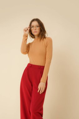 Stüdyo fotoğrafları. Sade güzel kıyafetler giyen, bej balıkçı yaka ve kırmızı geniş paça pantolon giyen genç bir manken.