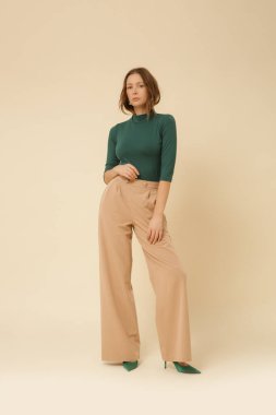 Stüdyo fotoğrafları, basit güzel kıyafetler, yeşil balıkçı yaka kazak ve bej geniş paça pantolon giyen genç bir model.