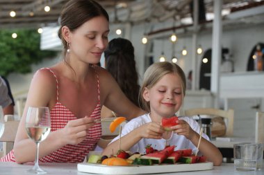 Annesi ve küçük tatlı kızı plaj barında sıcak bir yaz gününün tadını çıkarıyor, meyve tabağından meyve yiyorlar.