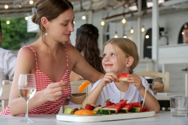 Annesi ve küçük tatlı kızı plaj barında sıcak bir yaz gününün tadını çıkarıyor, meyve tabağından meyve yiyorlar.