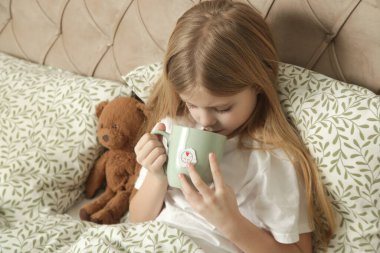 Hasta küçük kız bir fincan çayla yatakta, soğuk algınlığı ve grip mevsimi, doğal bitkisel tedavi