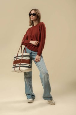 Klasik sonbahar kıyafetleri içindeki kadın fotoğrafları. Mavi kot, kahverengi kazak, güneş gözlüğü ve büyük çanta..