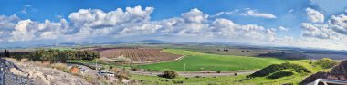 Nefes kesen panoramik bir manzara, geniş yeşil alanlar, ekilmiş tarım arazileri, ve dağınık bulutlu parlak mavi gökyüzünün altında yuvarlanan tepeler. Manzara, doğal güzelliği tarım cazibesiyle harmanlayarak ufuk çizgisine kadar uzanıyor..