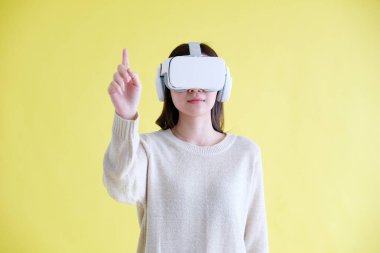 Sanal gerçeklik takan Asyalı bir kadın, sanal gerçeklik kulaklığı takıyor ve izole edilmiş sarı arka planda dururken bir şeye elle dokunmaya çalışıyor.