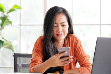 Evde çalışmak, evde dizüstü bilgisayarla çalışırken cep telefonu kullanan Asyalı bir kadın, internetten öğrenen mutlu bir kız, online eğitim.