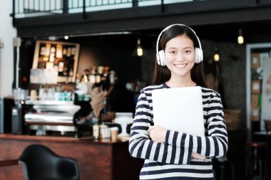 Kablosuz kulaklık takan ve elinde dizüstü bilgisayarla kafenin arka planında duran mutlu güzel kadın, insanlar ve teknoloji, yaşam tarzı