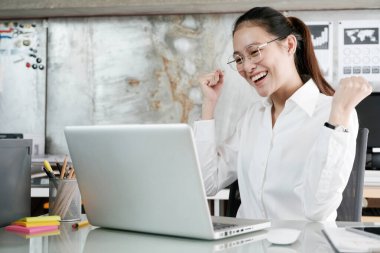 Ofis masasındaki bilgisayar ekranına bakarken kollarını kaldırıp gülümseyen genç bir iş kadını. İş konseptinde başarı.