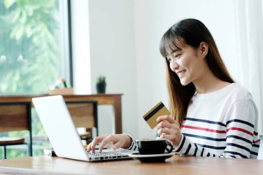 Kredi kartı tutan Asyalı genç bir kadın kafeterya, iş ve teknoloji konsepti, dijital pazarlama, günlük yaşam tarzı alışverişi yapmak için dizüstü bilgisayar kullanıyor.