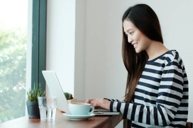Güzel Asyalı kadın pencere kenarında otururken dizüstü bilgisayar kullanıyor, evde çalışıyor, insanların eğitimi, işi ve teknolojisi, günlük yaşam tarzı kavramı
