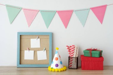 Mavi antika ahşap çerçevenin üzerinde asılı renkli parti bayrakları boş kağıt kartlar ve hediye kutusu, beyaz duvarda parti süsleri, doğum günü, yıldönümü, kutlama etkinliği, festival geçmişi