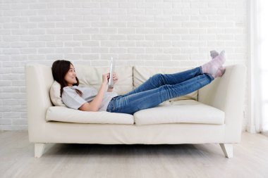 Genç Asyalı kadın gülümseyen, mutlu ve rahat bir tablet kullanıyor beyaz oda arka planında koltukta uzanırken, insanlar ve teknoloji, yaşam tarzı