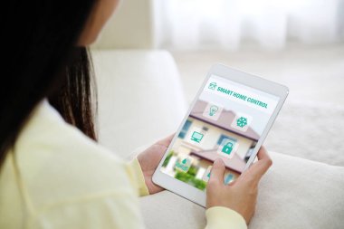 Evde kanepede otururken ekranda akıllı ev denetimi simgesi olan tablet kullanan kadın, akıllı ev kontrol kavramı