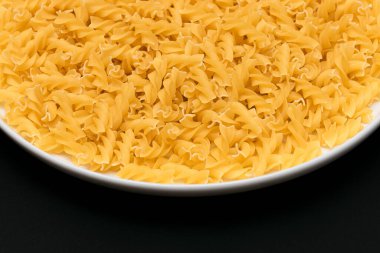 Pişmemiş Fusilli Makarna Siyah Arkaplanda Beyaz Tabakta Yatıyor. Çiğ ve kuru makarna. Sağlıksız ve şişman yiyecekler