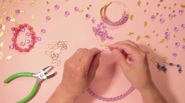 El yapımı Mücevher. Beyaz Tişörtlü Beyaz Avrupalı Kadın Pembe Masa 'da El yapımı Bijouterie, Boncuk ve Bilezik Yapıyor. Sanatçılar İş Yeri - Üst Görünüm, Sabit Çekim