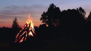 Big Burning Campfire at Summer Evening against the Blue Sky. Wood on Fire. Flying Sparks. Travel and Tourism Concept. Giant Flaming Bonfire - Static Shot, Timelapse