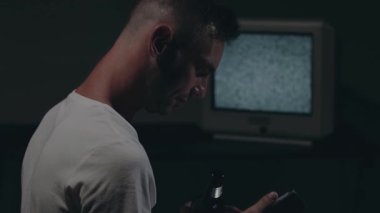 Beyaz tişörtlü adam Karanlık Oda 'da Eski Gümüş TV' ye Karşı Oturuyor. Bira içip, akıllı telefonunu kullanmaktan başka. Televizyon sinyalini ara. Ekrandaki bozulma. Arka Görünüm, Orta Yakın Çekim, Film Tanesi